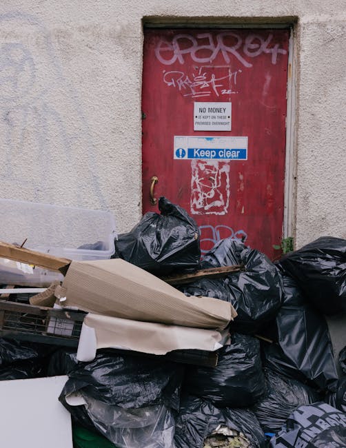 A red metal door with graffiti tags and a white sign that reads 'No Money, Keep Clear' is situated within an exterior wall of a building with a textured, off-white finish. In front of the door, there 