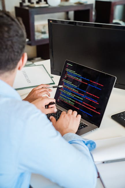 A person with short dark hair, wearing a light blue shirt, is seated at a desk working on a laptop with a dark background screen displaying colourful lines of programming code. The individual is holdi